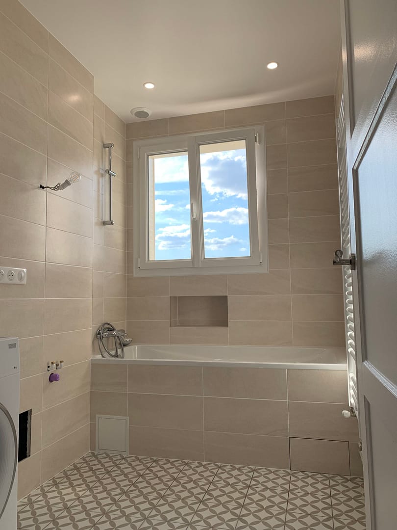 "Salle de bain moderne rénovée avec carrelage beige, baignoire blanche, et fenêtre ouverte sur le ciel parisien"